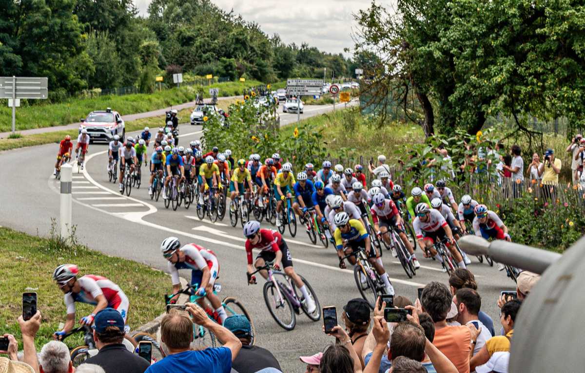 Peloton clustering at the start line with team buses and race officials in the background